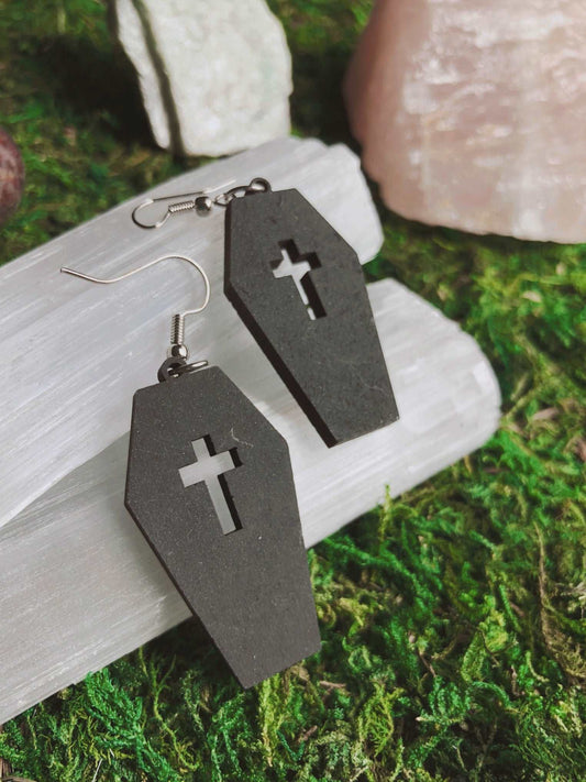 Pictured is a pair of earrings featuring a black coffin with a cut out cross on top. They are made of wood. 