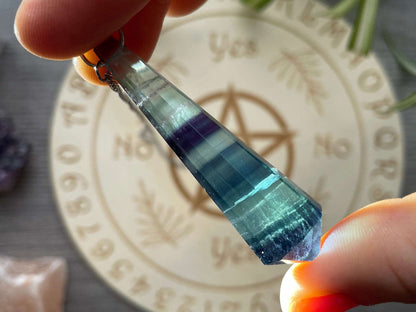 A blue and purple fluorite crystal pendulum sits atop a wooden divination board.