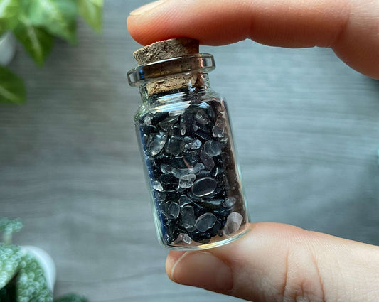 Pictured is a small glass vial with a cork stopper. Inside the jar are black tourmaline and clear quartz chips.