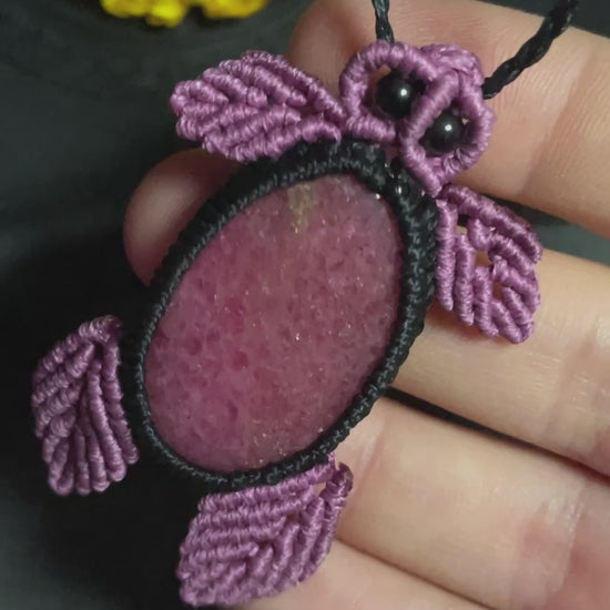 Pictured is a rhodonite cabochon wrapped in macrame thread. The rhodonite pendant is in the shape of a sea turtle. A gothic book and flowers are nearby.