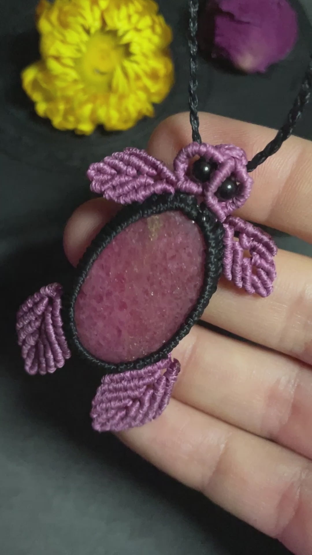 Pictured is a rhodonite cabochon wrapped in macrame thread. The rhodonite pendant is in the shape of a sea turtle. A gothic book and flowers are nearby.