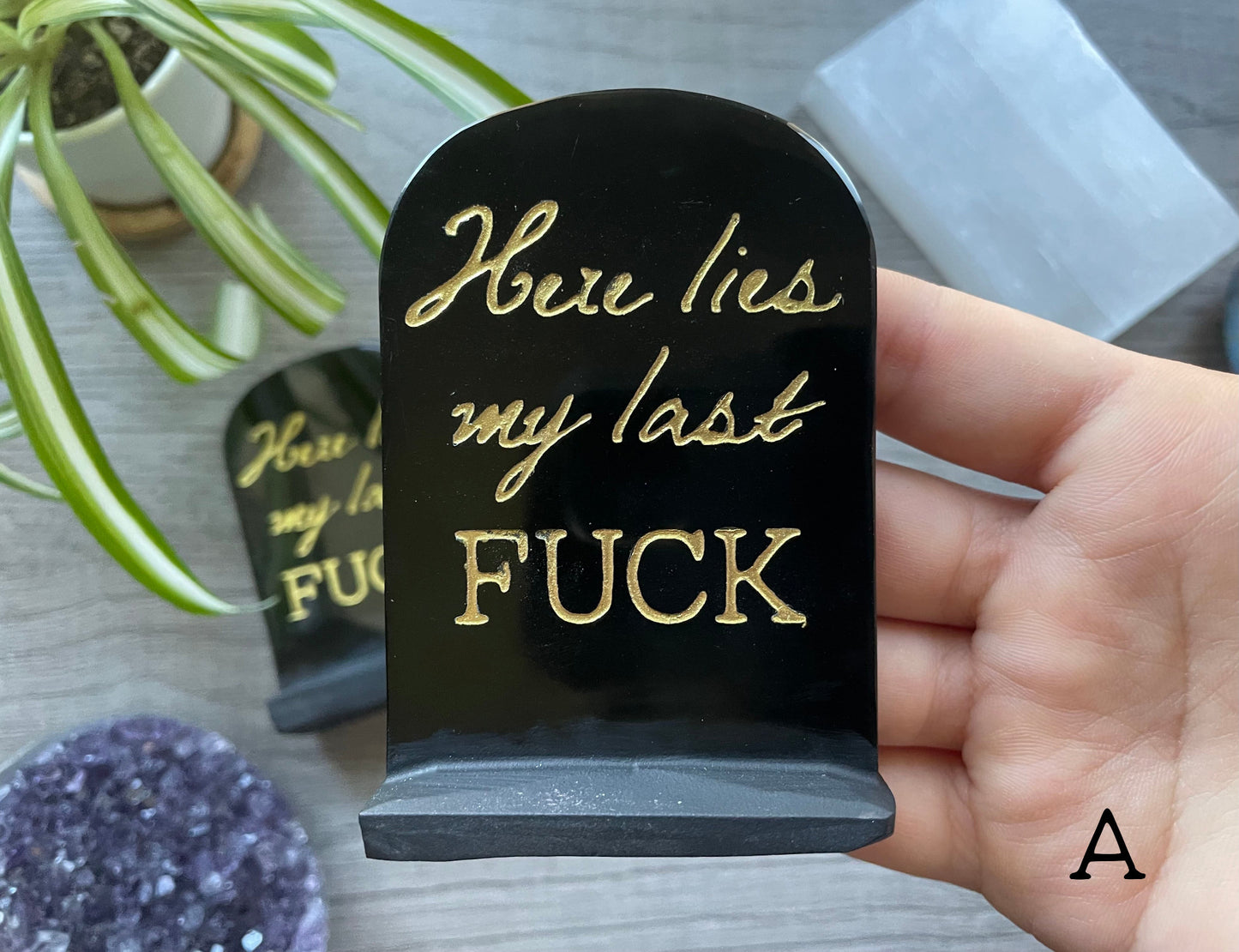 An image of a carved obsidian tombstone with the words "Here Lies My Last Fuck" written in gold letters on it. The tombstone is a humorous and cheeky statement piece that adds a touch of personality and edge to any space, and serves as a reminder to let go of negativity and embrace a more carefree and lighthearted attitude.