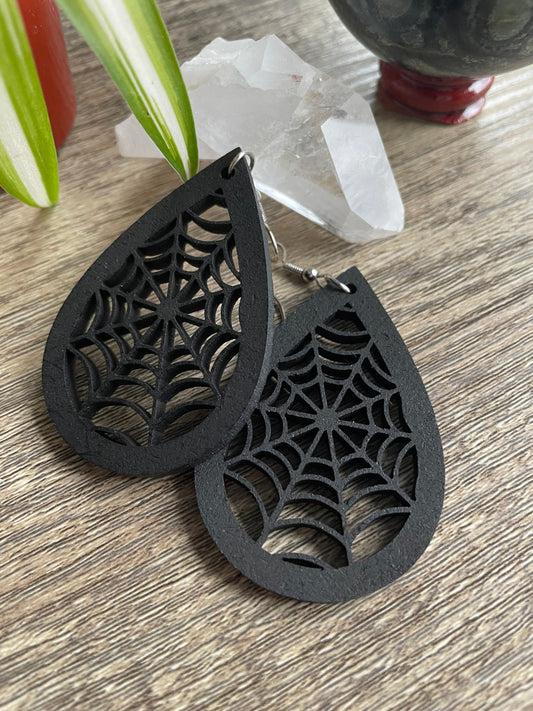 Pictured is a pair of earrings featuring black teardrop-shaped spider webs. They are made of wood. 