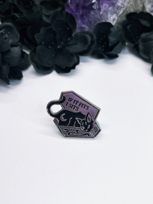 An image of a silver-colored enamel pin featuring a black cat sitting in a coffin with the words "If It Fits, I Sits" written in black letters. The design features a black cat sitting comfortably inside a black coffin, with its eyes closed and a content expression on its face.