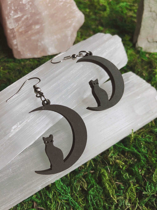 Pictured is a pair of earrings featuring a black cat sitting on a black crescent moon. They are made of wood. 
