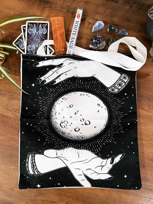 Pictured is a large canvas tote bag featuring an image of two hands around an image of a moon.