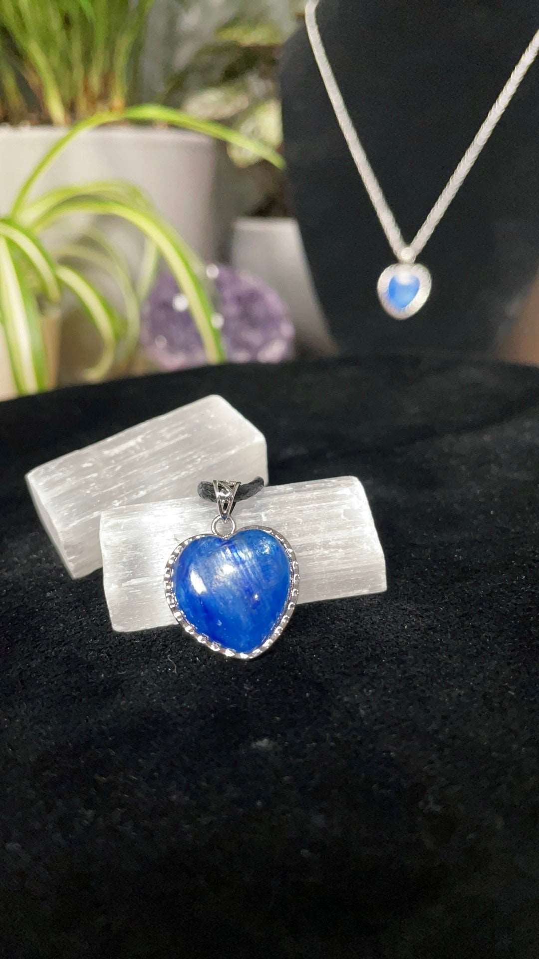 An image of a blue kyanite heart shaped silver pendant on a necklace. It sits atop some selenite chunks and a black velvet surface.