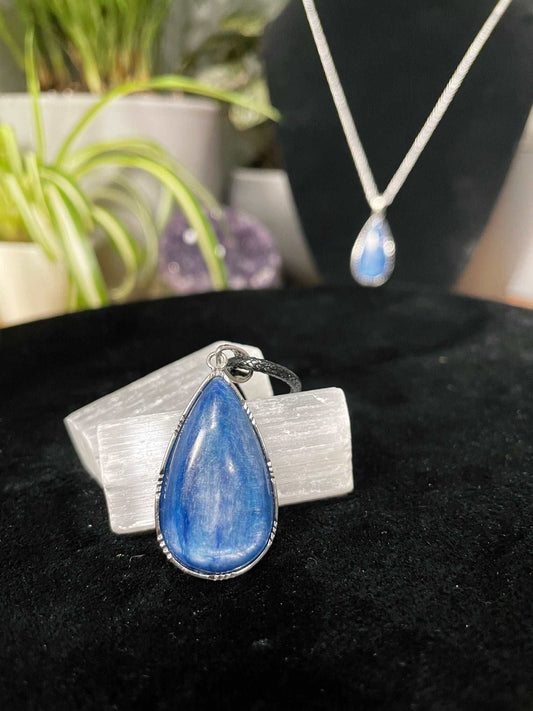 An image of a blue kyanite teardrop shaped silver pendant on a necklace. It sits atop some selenite chunks and a black velvet surface.