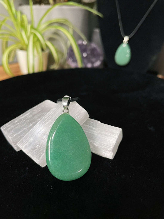 An image of a green aventurine teardrop shaped pendant on a necklace. It sits atop some selenite chunks and a black velvet surface.