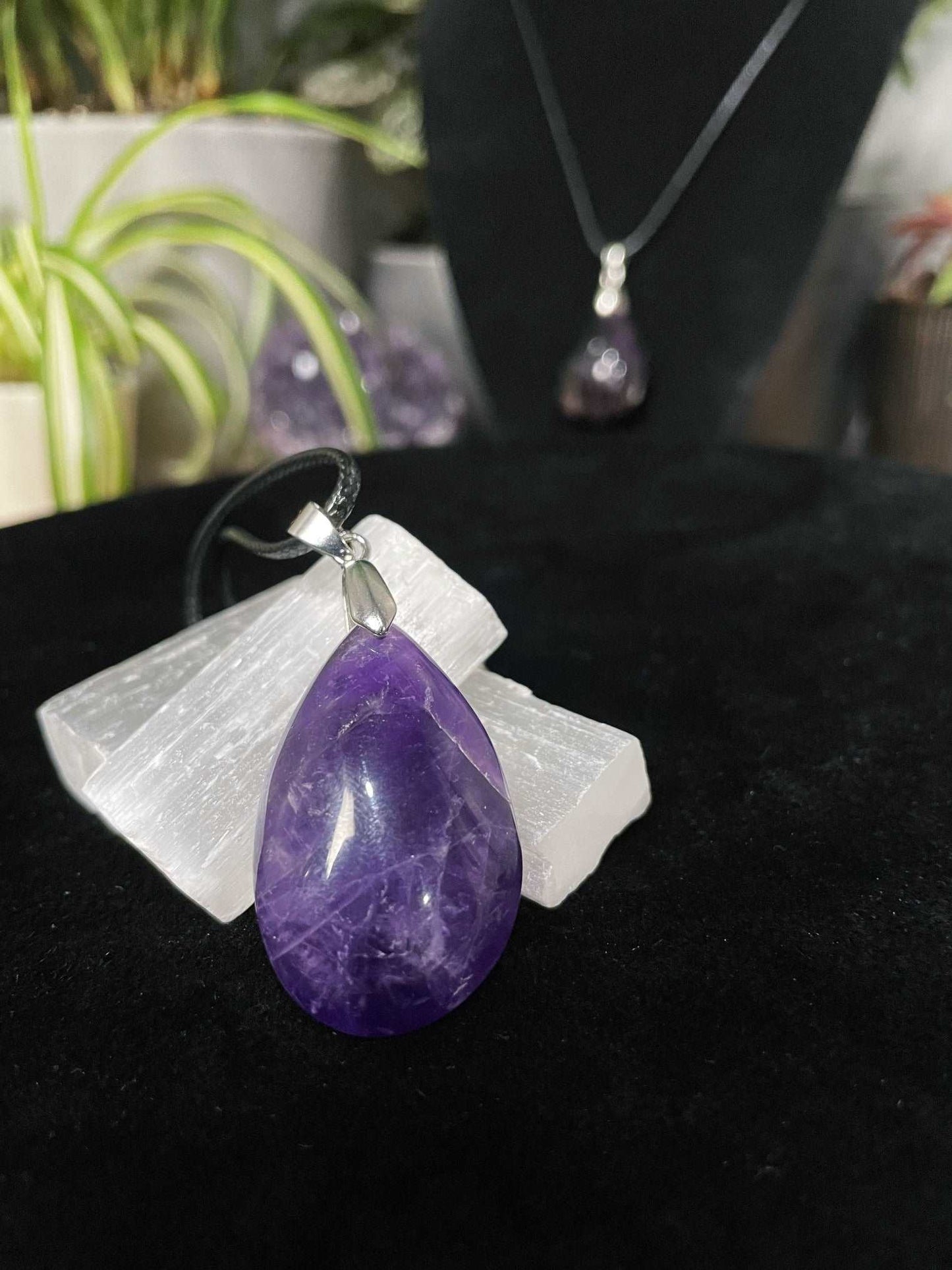 An image of a purple amethyst teardrop shaped pendant on a necklace. It sits atop some selenite chunks and a black velvet surface.