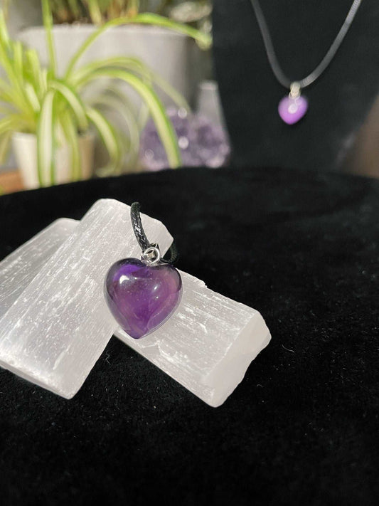 An image of a purple amethyst heart shaped pendant on a necklace. It sits atop some selenite chunks and a black velvet surface.
