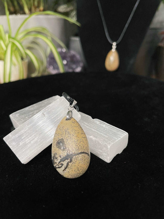 An image of a Chinese painting stone teardrop shaped pendant on a necklace. It sits atop some selenite chunks and a black velvet surface.