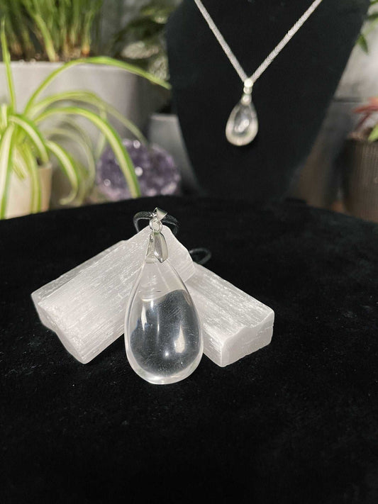 An image of a clear quartz teardrop shaped pendant on a necklace. It sits atop some selenite chunks and a black velvet surface.