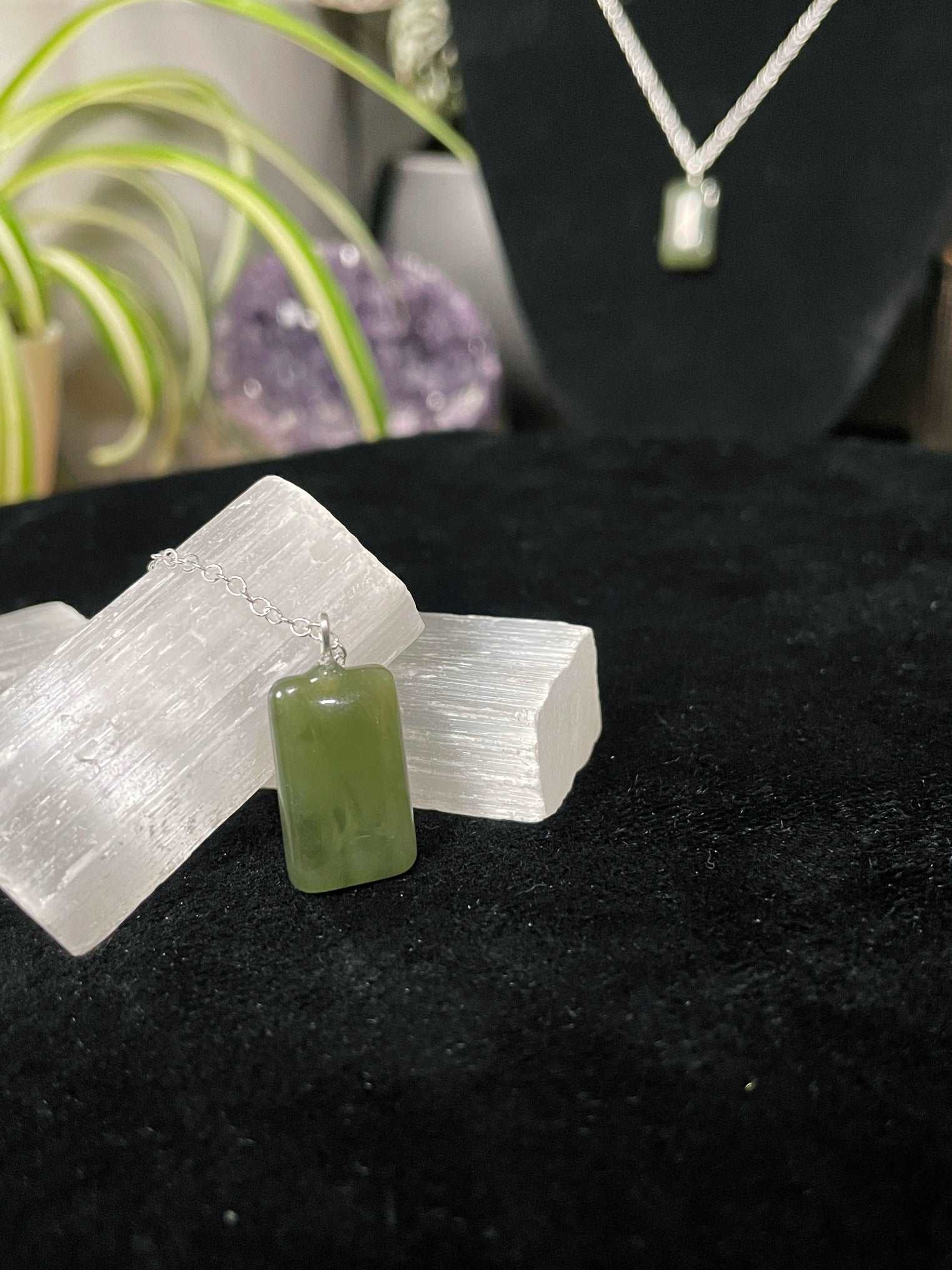 An image of a green nephrite jade rectangle shaped pendant on a necklace. It sits atop some selenite chunks and a black velvet surface.