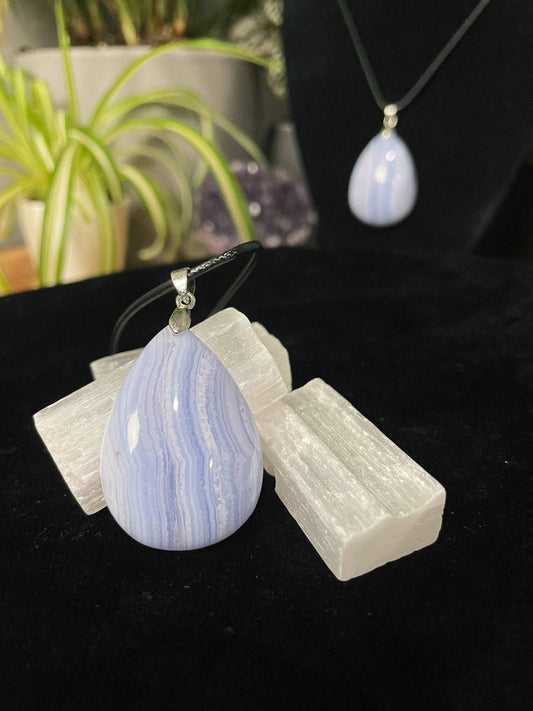 An image of a blue lace agate teardrop shaped pendant on a necklace. It sits atop some selenite chunks and a black velvet surface.