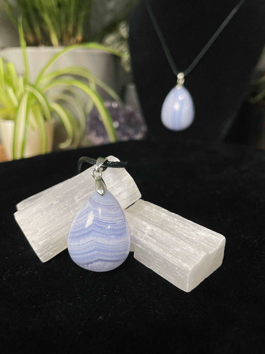 An image of a blue lace agate teardrop shaped pendant on a necklace. It sits atop some selenite chunks and a black velvet surface.