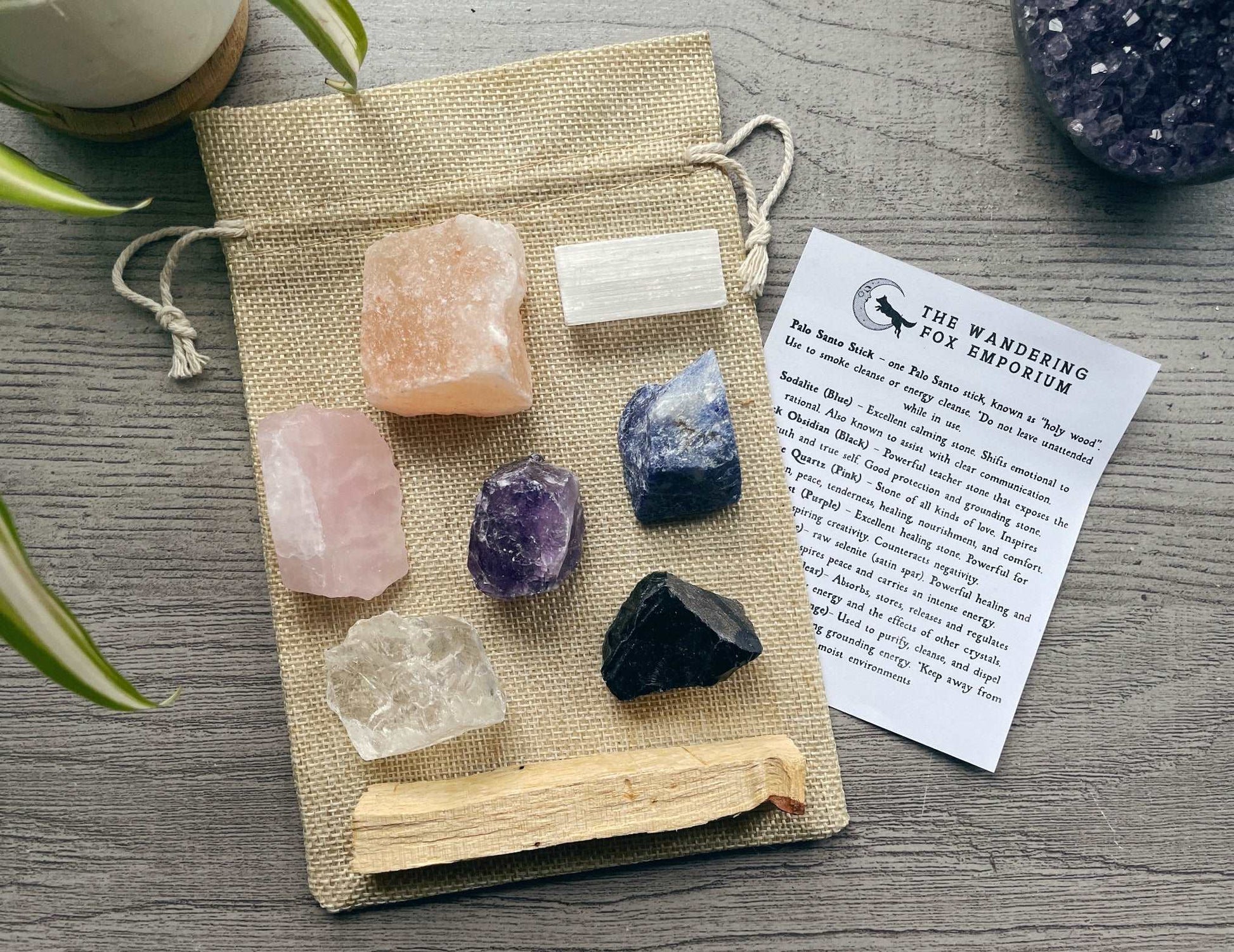 Pictured is a crystal starter set with various raw crystals and a palo santo stick. A piece of paper is nearby with the information about the crystals on it.
