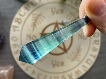 A blue and purple fluorite crystal pendulum sits atop a wooden divination board.