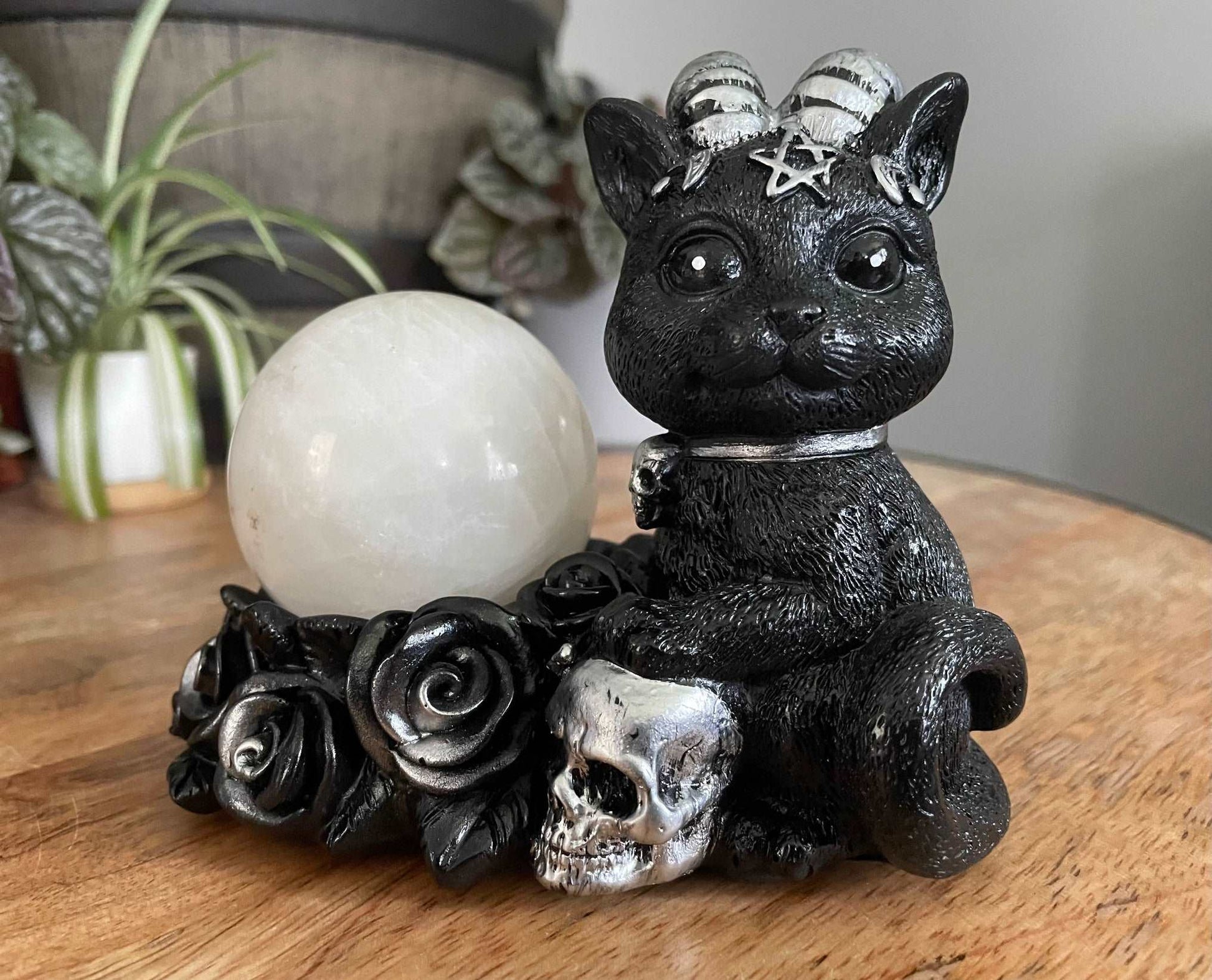 Pictured is a sphere stand of a black cat with horns sitting next to some silver and black roses and a silver skull.