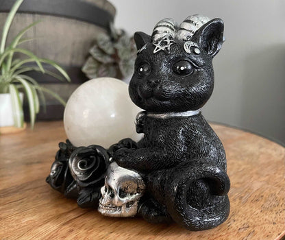 Pictured is a sphere stand of a black cat with horns sitting next to some silver and black roses and a silver skull.