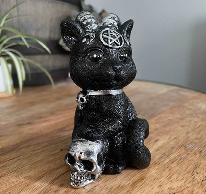 Pictured is a resin statue of a black cat with horns and a pentagram on its forehead with a paw on a silver skull.