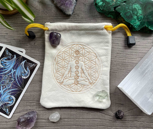Pictured is a cream-coloured, faux-suede drawstring bag with a flower of life and a meditating person with labelled chakras printed on the front.