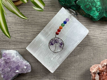 A tree of life with amethyst crystals as the leaves on the tree is in the image. The keychain has a number of small multi-coloured beads to represent the chakras on the chain. The keychain sits atop a slab of selenite. Malachite, pink amethyst, and amethyst are nearby.