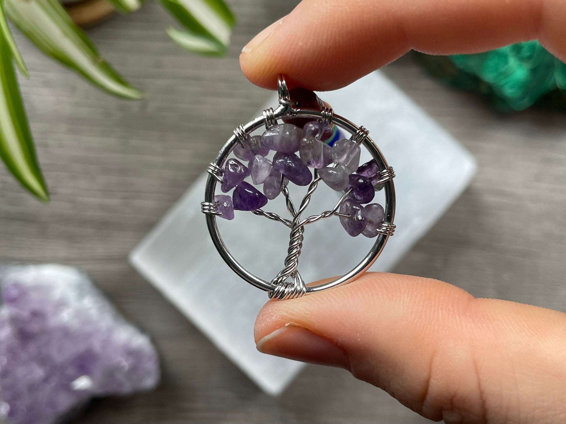 A tree of life with amethyst crystals as the leaves on the tree is in the image. The keychain has a number of small multi-coloured beads to represent the chakras on the chain. The keychain sits atop a slab of selenite. Malachite, pink amethyst, and amethyst are nearby.