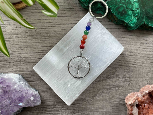 A tree of life with quartz crystals as the leaves on the tree is in the image. The keychain has a number of small multi-coloured beads to represent the chakras on the chain. The keychain sits atop a slab of selenite. Malachite, pink amethyst, and amethyst are nearby.
