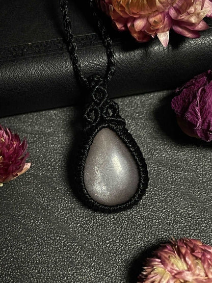 Pictured is a grey moonstone cabochon wrapped in macrame thread. A gothic book and flowers are nearby.