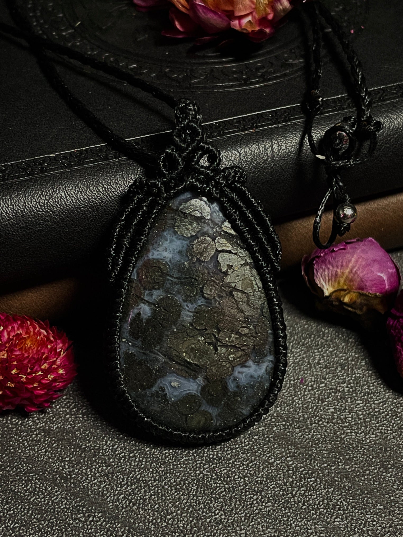 Pictured is a marcasite in agate cabochon wrapped in macrame thread. A gothic book and flowers are nearby.