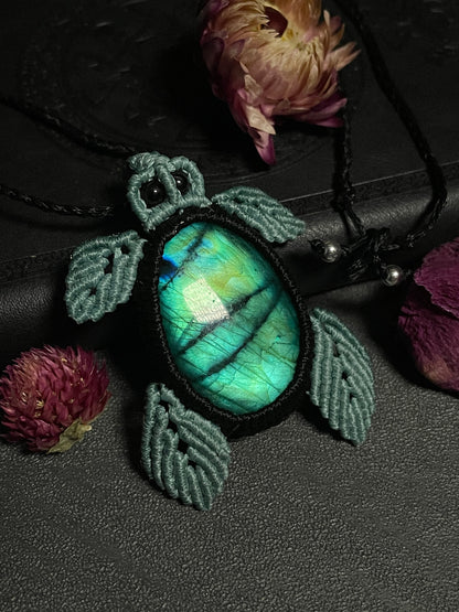 Pictured is a macrame pendant shaped like a sea turtle. Its shell is a labradorite cabochon with blue and green flash. Its eyes are black tourmaline beads.