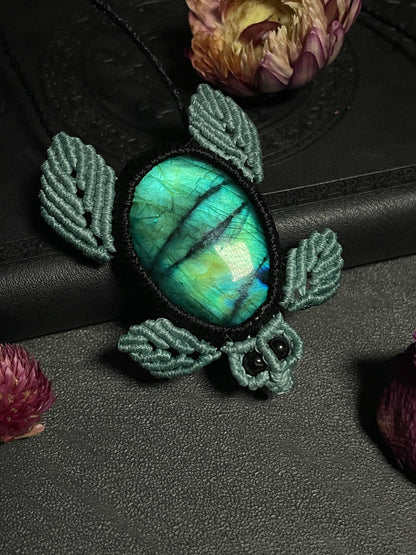 Pictured is a macrame pendant shaped like a sea turtle. Its shell is a labradorite cabochon with blue and green flash. Its eyes are black tourmaline beads.