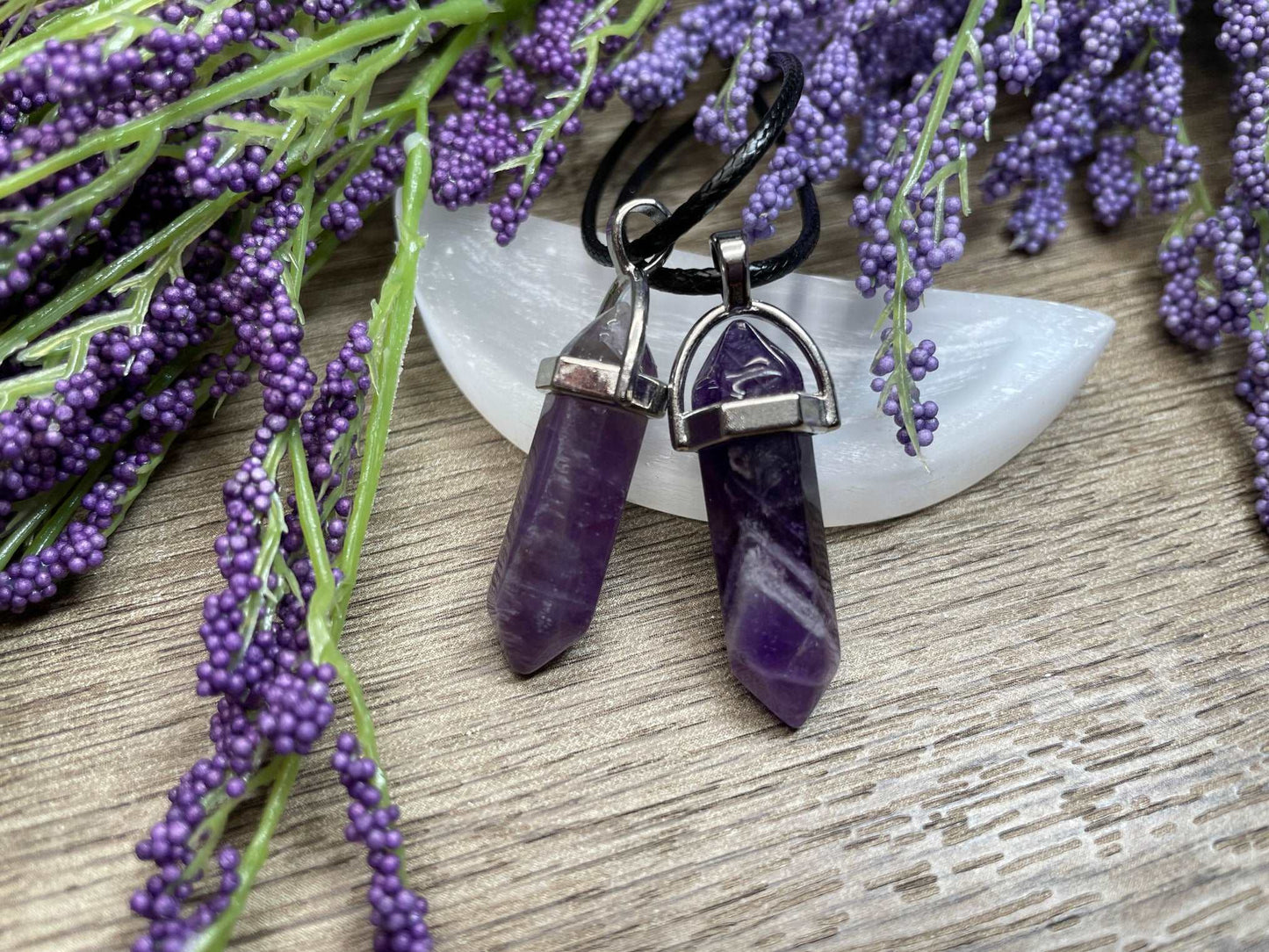 An image of purple amethyst double-terminated polished crystal necklaces sitting on selenite and surrounded by lavender on a wood surface..