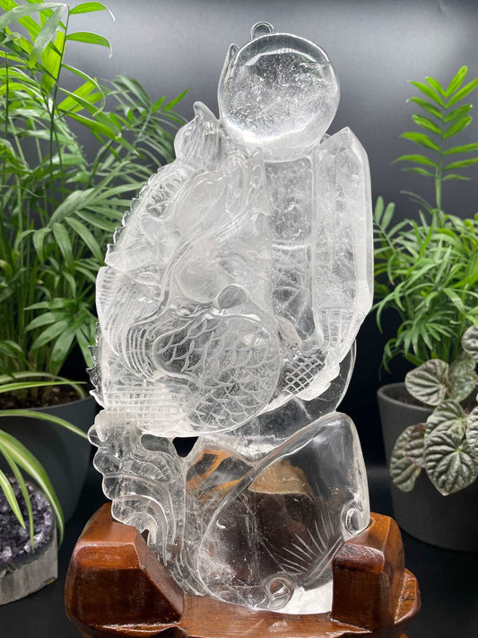 Pictured is a gorgeous clear quartz carving of a dragon holding a crystal wand and chasing a lucky sphere. It stands on a custom, hand-carved wood stand.