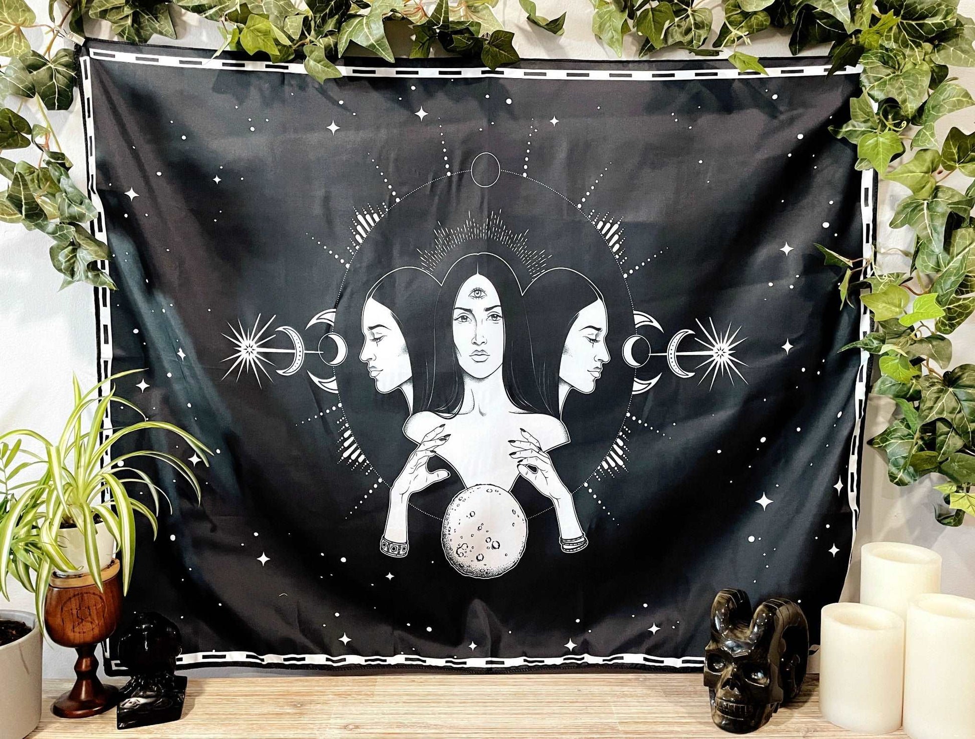 Pictured is a large wall tapestry with a black background and three women representing three goddesses or Hecate printed in white on it.