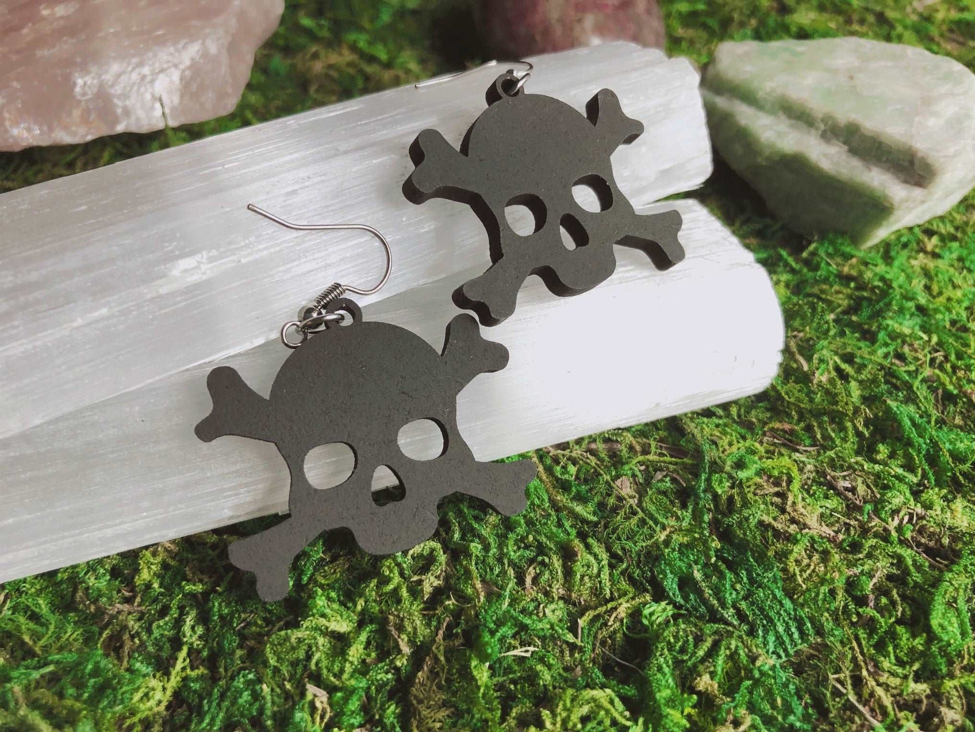 Pictured is a pair of earrings featuring a black skull and crossbone. They are made of wood. 