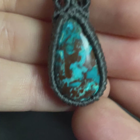 Pictured is a chrysocolla and cuprite cabochon wrapped in macrame thread. A gothic book and flowers are nearby.