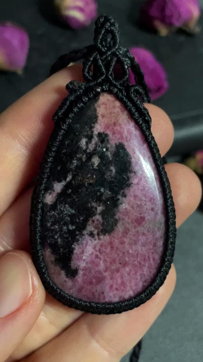 Pictured is a rhodonite cabochon wrapped in macrame thread. A gothic book and flowers are nearby.