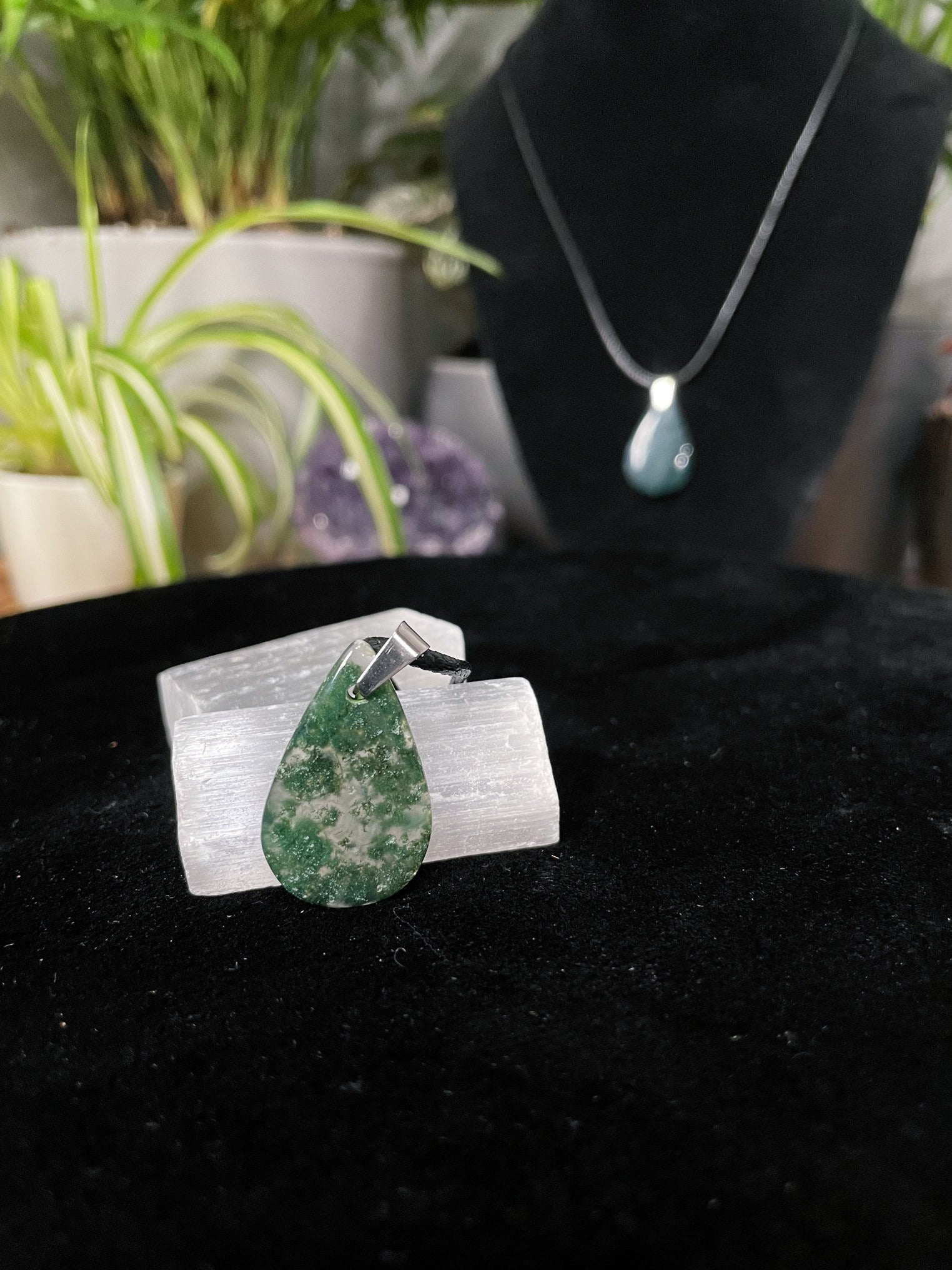 An image of a green moss agate teardrop shaped pendant on a necklace. It sits atop some selenite chunks and a black velvet surface.