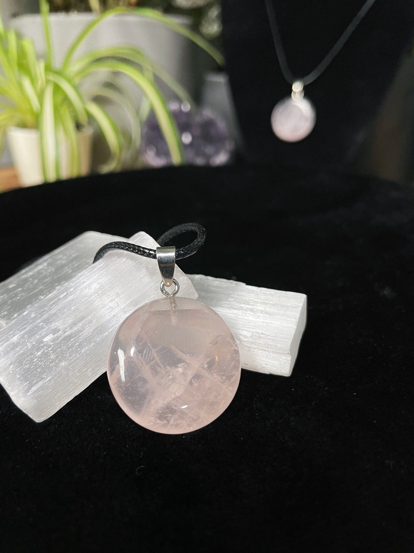 An image of a light pink rose quartz circle shaped pendant on a necklace. It sits atop some selenite chunks and a black velvet surface.
