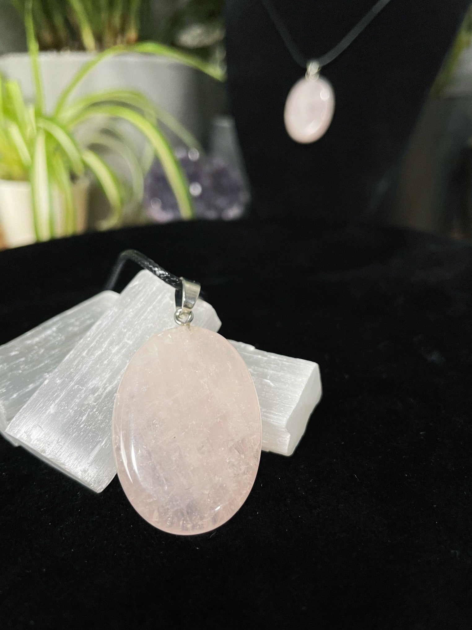 An image of a pink rose quartz oval shaped pendant on a necklace. It sits atop some selenite chunks and a black velvet surface.