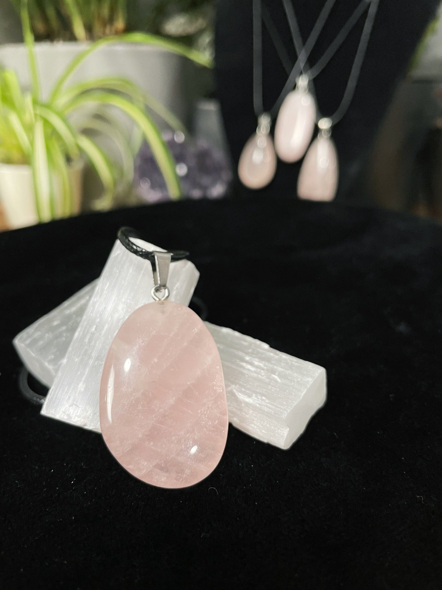 An image of a pink rose quartz random shaped pendant on a necklace. It sits atop some selenite chunks and a black velvet surface.