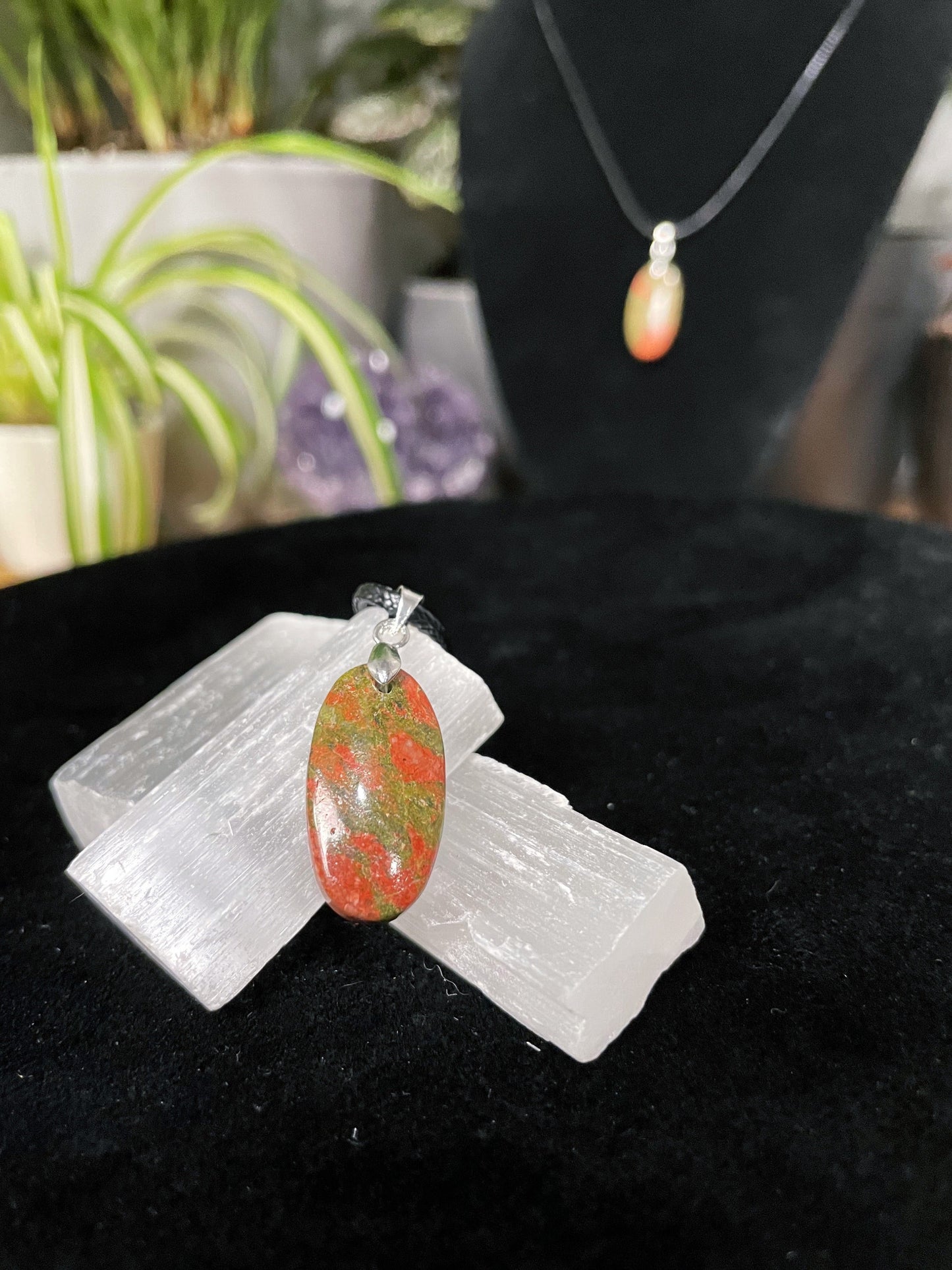 An image of an unakite oval shaped pendant on a necklace. It sits atop some selenite chunks and a black velvet surface.