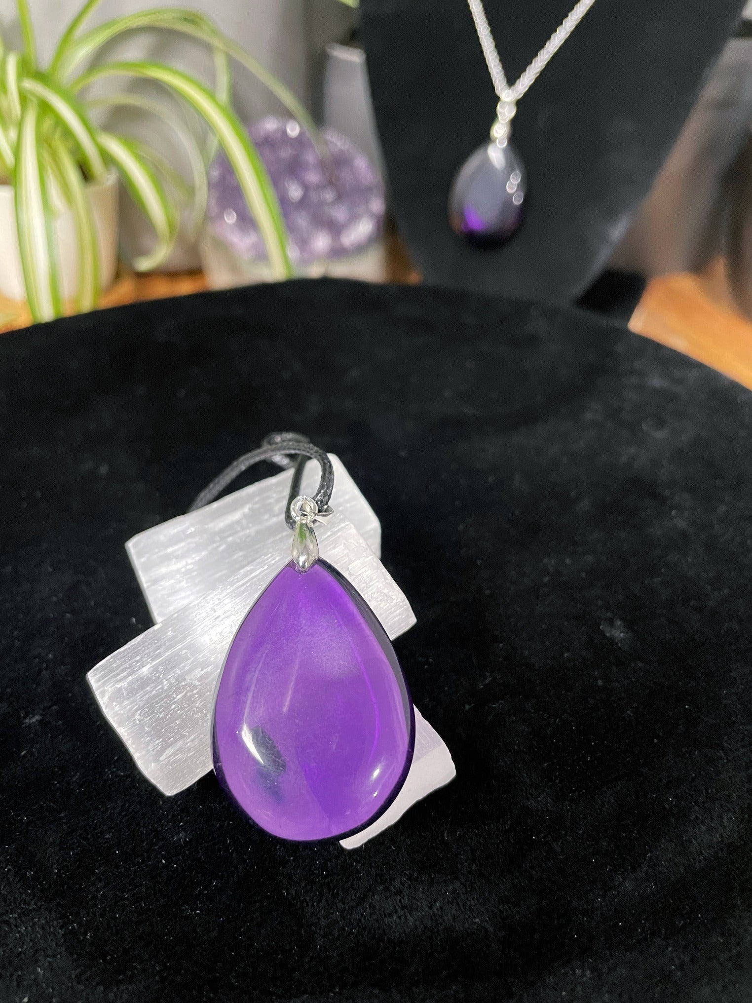 An image of a high-quality purple amethyst teardrop shaped pendant on a necklace. It sits atop some selenite chunks and a black velvet surface.