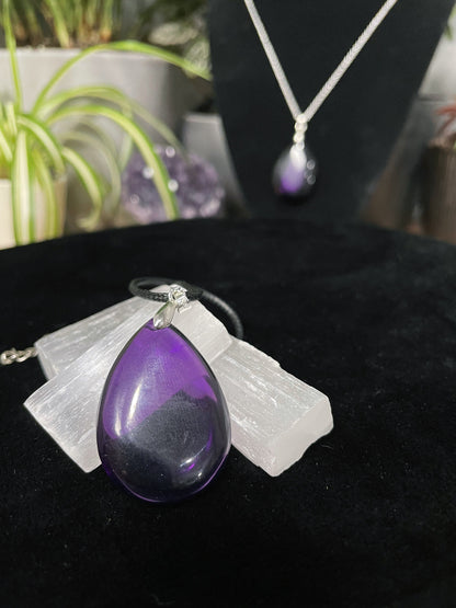 An image of a high-quality purple amethyst teardrop shaped pendant on a necklace. It sits atop some selenite chunks and a black velvet surface.