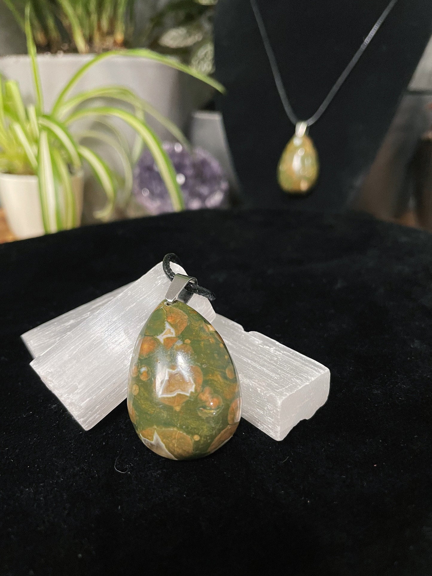An image of a multicoloured rainforest jasper teardrop shaped pendant on a necklace. It sits atop some selenite chunks and a black velvet surface.