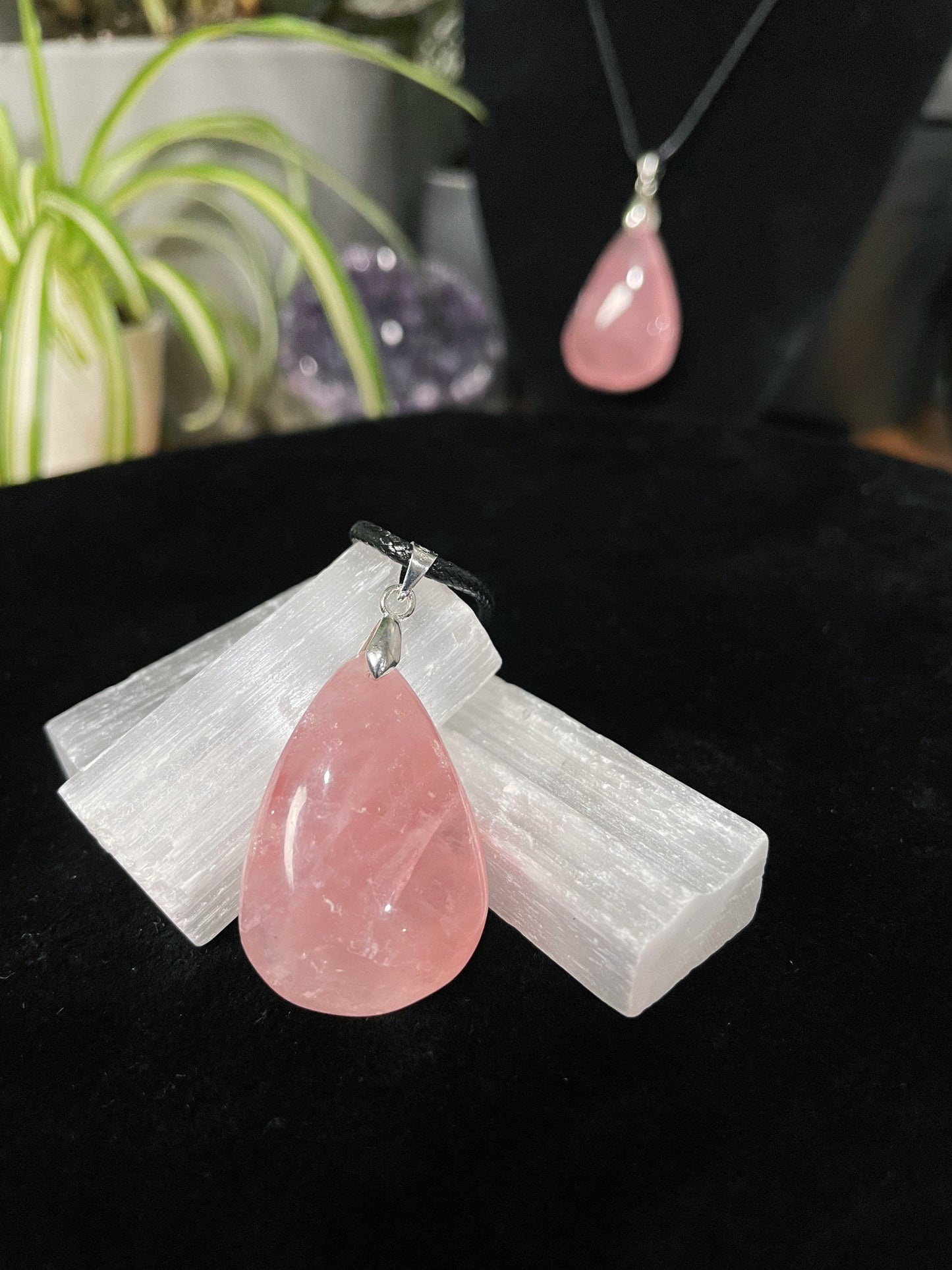 An image of a pink rose quartz teardrop shaped pendant on a necklace. It sits atop some selenite chunks and a black velvet surface.