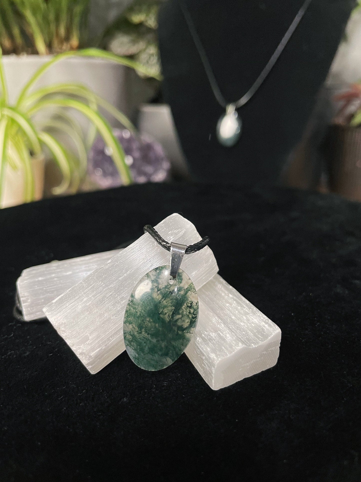 An image of a green moss agate oval shaped pendant on a necklace. It sits atop some selenite chunks and a black velvet surface.