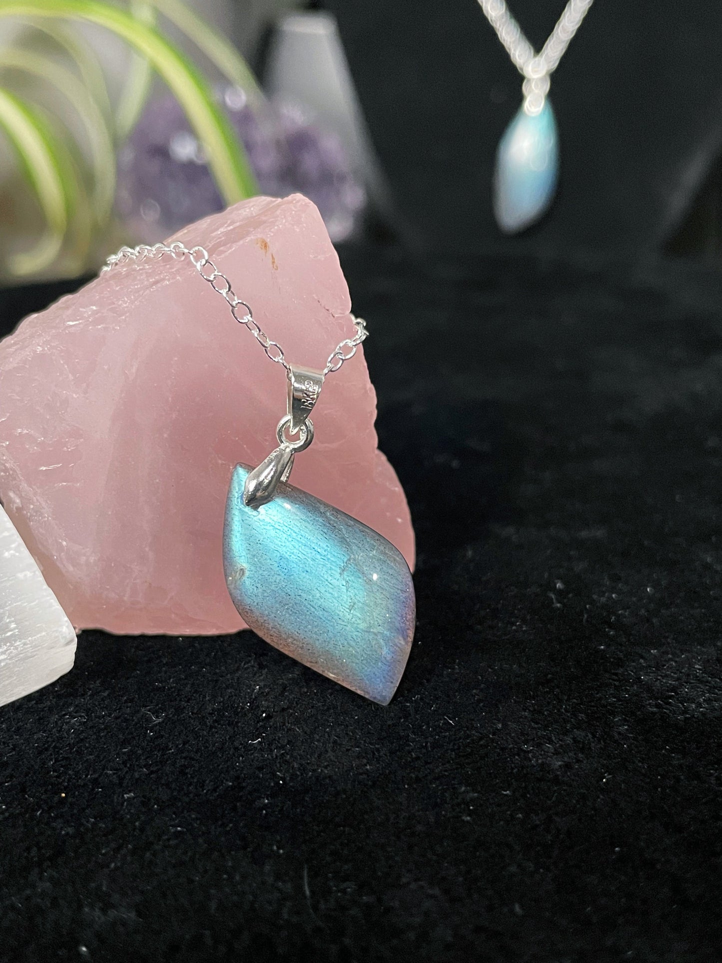 An image of a high-quality blue flash labradorite leaf shaped pendant on a necklace. It sits atop some selenite chunks and a black velvet surface.